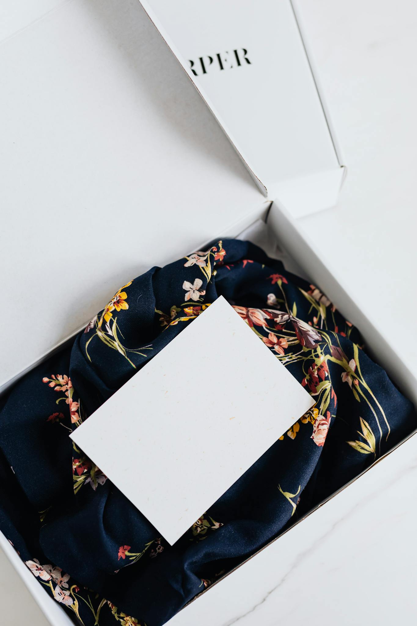 From above of stylish black silk floral pattern cloth with white visit card mockup placed in white carton box after receiving postal delivery of online order